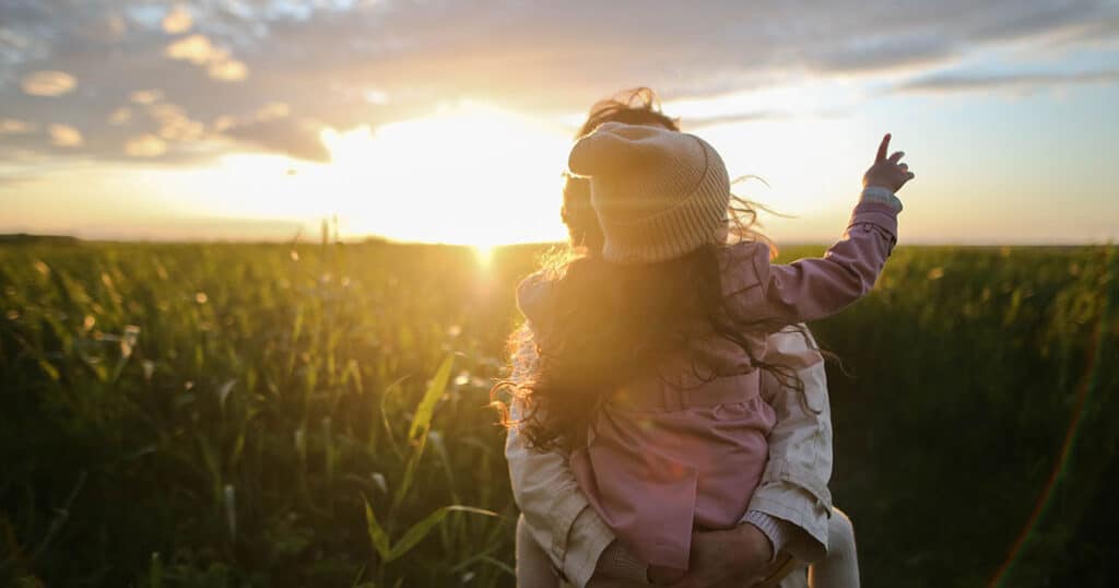 A mother holding her child in a field at sunset, both facing away from the camera.