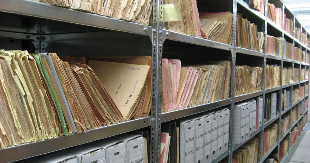 Rows of metal shelves filled with stacks of old, worn-out files and folders in an archive room.