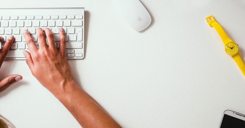 The image shows a person typing on a white keyboard. A white mouse and a yellow watch are also visible on the desk.