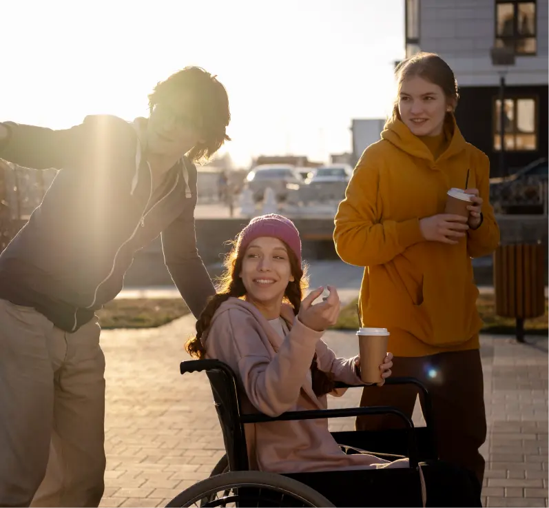 Three friends, two women holding coffee cups, with one seated in a wheelchair being pushed by a male friend who is pointing at something, all looking in the same direction.