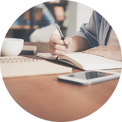 A person's hand holding a pen, writing on paper at a table with books and a phone nearby.